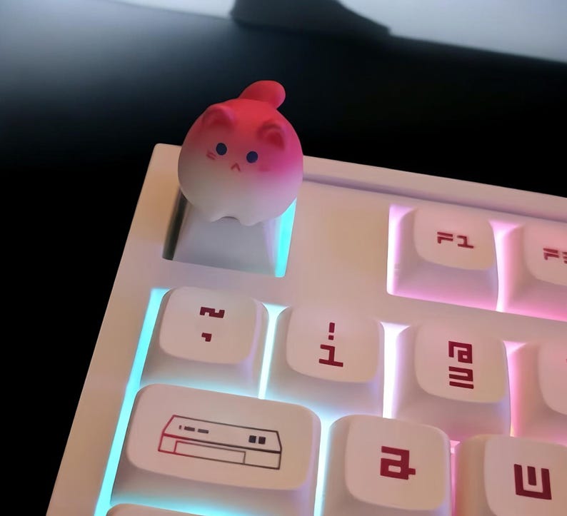 May include: A white keyboard with pink and blue backlighting. A pink and white cat-shaped keycap is on the 'F' key.