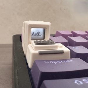 May include: A white computer-shaped keycap with a miniature keyboard and the word "hello" on the screen. The keycap is on a keyboard with purple keys, including one labeled "Capslock".