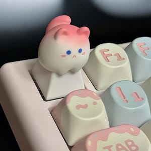 May include: A pink and white cat-shaped keycap with blue eyes sits on a white keycap. The keycap is on a pink and white keyboard with other keys visible, including keys labeled "F1", "!", and "TAB".