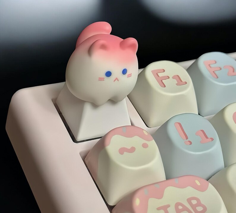 May include: A pink and white cat-shaped keycap with blue eyes sits on a white keycap. The keycap is on a pink and white keyboard with other keys visible, including keys labeled "F1", "!", and "TAB".