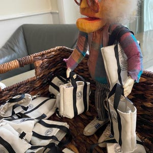May include: A collection of small, white canvas tote bags with black straps and a black logo. The bags are arranged in a wicker basket. A puppet wearing a blue and red plaid shirt and a blue cap is holding one of the bags.