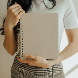 May include: A person holding a gray spiral bound journal with the text "Plan Home" embossed on the front cover.