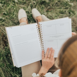 May include: A person sitting in a grassy field, holding open a spiral bound journal with lined pages and a section with prompts for writing. The journal has a beige cover with a floral pattern.