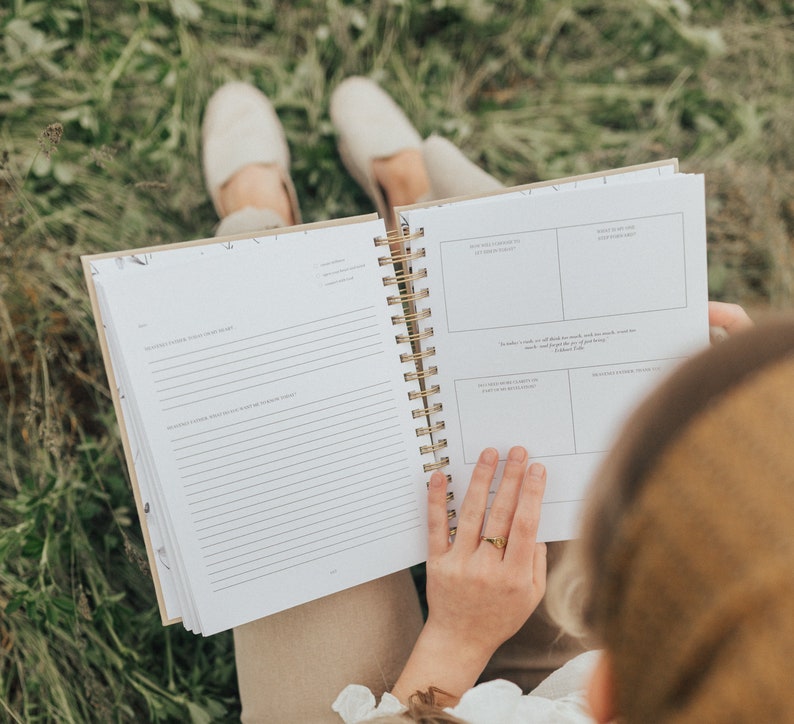 May include: A person sitting in a grassy field, holding open a spiral bound journal with lined pages and a section with prompts for writing. The journal has a beige cover with a floral pattern.