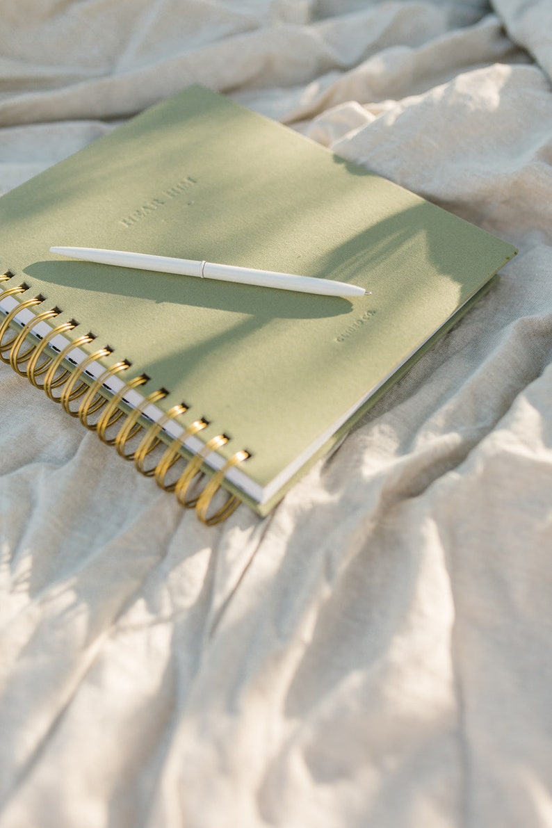 May include: A green spiral bound journal with gold rings and a white pen lying on top. The journal has the words "Dear Diary" embossed on the front cover.
