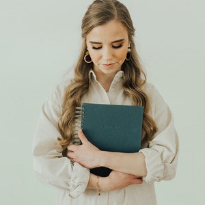 May include: A woman in a white shirt dress holds a blue spiral bound journal with the text "Dear Muse" on the cover.
