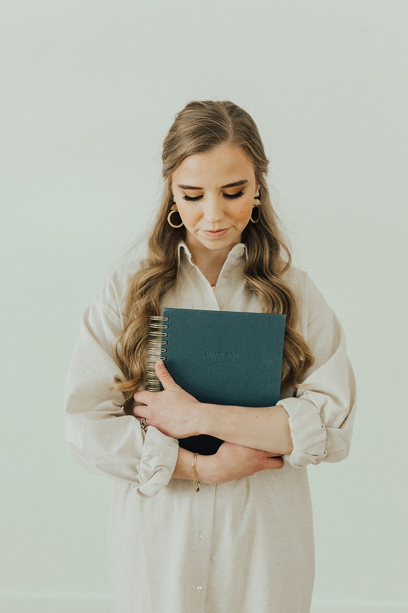 May include: A woman in a white shirt dress holds a blue spiral bound journal with the text "Dear Muse" on the cover.