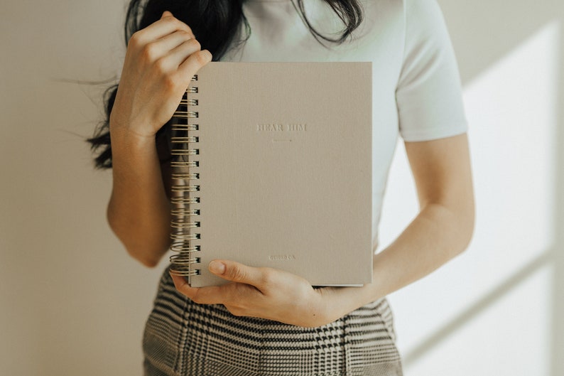 May include: A person holding a gray spiral bound journal with the text "Plan Home" embossed on the front cover.