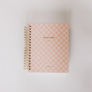 May include: A pink and white checkered notebook with a gold spiral binding. The cover has the text "HEAR HIM" in gold lettering and the text "CHARTER CO." in smaller gold lettering.