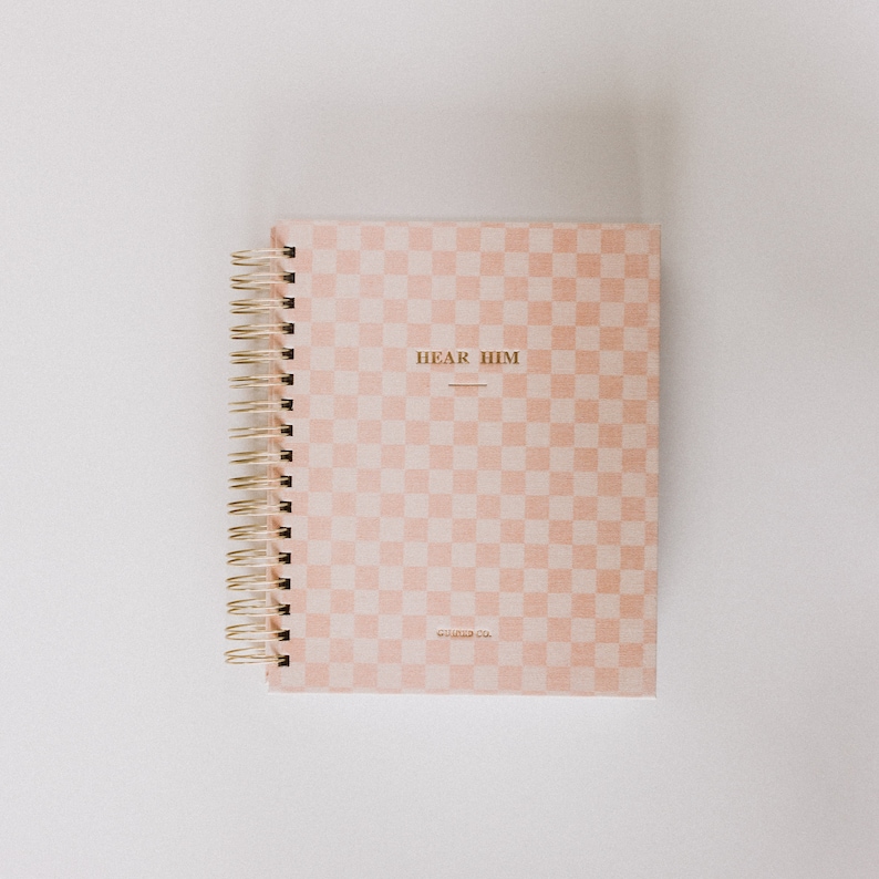 May include: A pink and white checkered notebook with a gold spiral binding. The cover has the text "HEAR HIM" in gold lettering and the text "CHARTER CO." in smaller gold lettering.