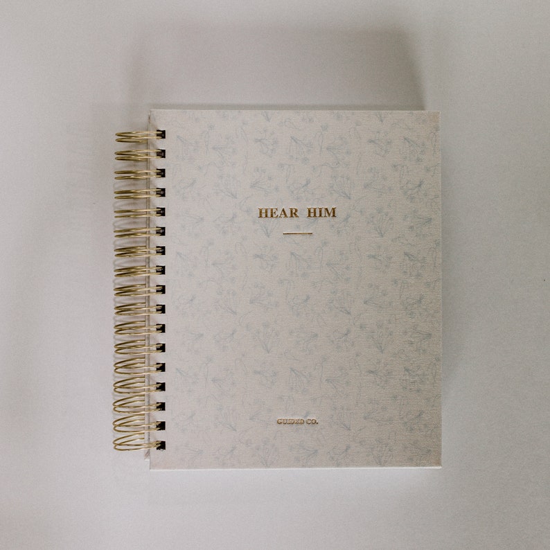 May include: A white spiral bound journal with a floral pattern and gold lettering. The cover reads "Hear Him" and "Guided Co."