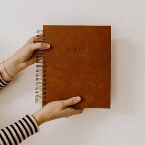 May include: A brown leather-bound journal with gold spiral binding. The cover has the words "Hear Him" embossed in gold.