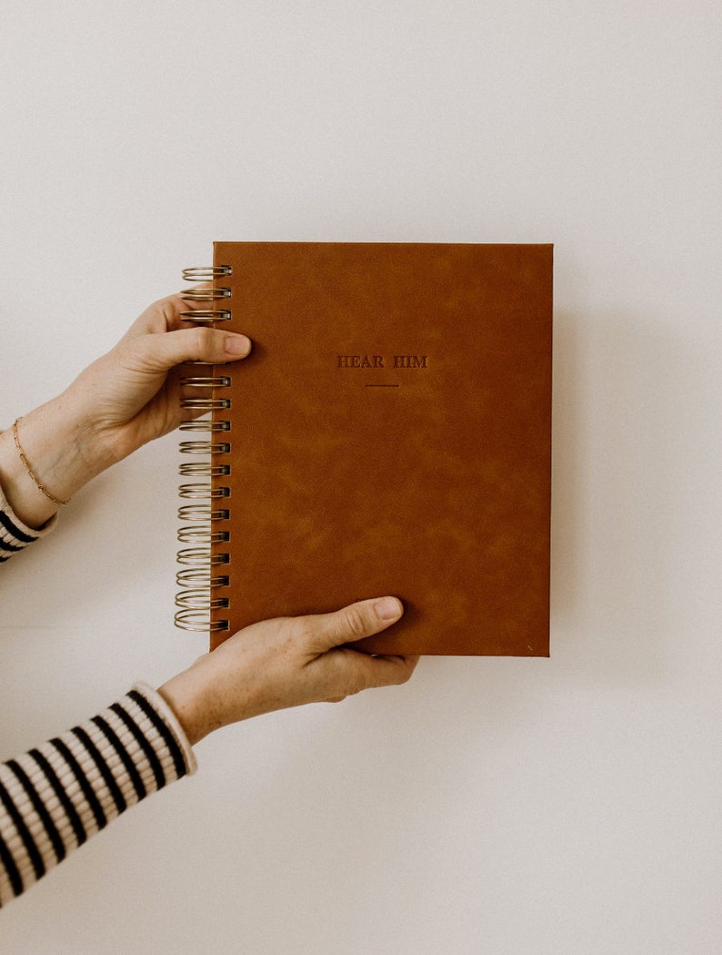 May include: A brown leather-bound journal with gold spiral binding. The cover has the words "Hear Him" embossed in gold.
