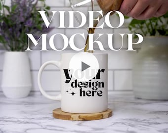 May include: White ceramic mug with a black play button and the text "your design here" on it. Coffee is being poured into the mug. The mug is on a wooden coaster. The text "VIDEO MOCKUP" is in the background.