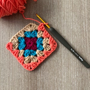 Puede incluir: Un cuadrado tejido a crochet con un centro colorido en tonos turquesa, burdeos y beige. El cuadrado est&aacute; rodeado por un borde de hilo de color coral. Un gancho de crochet negro con una punta dorada y el texto "5/0 3.00mm Tulip" est&aacute; sobre una superficie de madera.