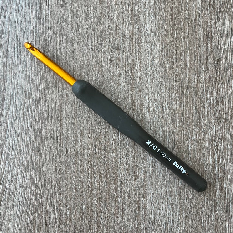 Puede incluir: Un ganchillo de oro y negro con mango negro y el texto "8/0 5.00mm Tulip" impreso en el lateral.