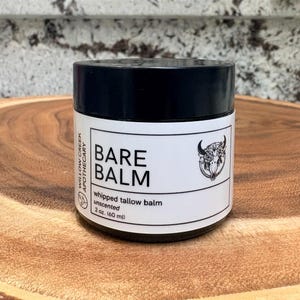May include: A black-lidded jar of Bare Balm, unscented whipped tallow balm, 60 ml. The label features the text "BARE BALM" and a floral skull illustration. The jar is on a wooden surface.