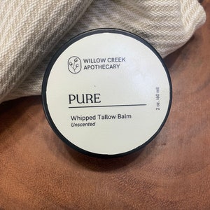 Peut inclure: Un pot rond noir avec une étiquette blanche indiquant "PURE" et "Whipped Tallow Balm Unscented". L'étiquette affiche également le logo Willow Creek Apothecary et indique 60 ml. Le pot est posé sur une surface en bois.