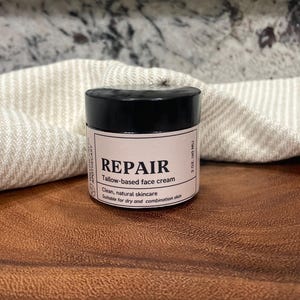 May include: A small, round jar of face cream with a black lid and a light pink label. The label reads "REPAIR" and "Tallow-based face cream." The jar is 60 ml and is suitable for dry and combination skin. The jar sits on a wooden surface.