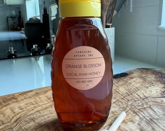 May include: A glass bottle of orange blossom honey with a yellow lid. The bottle is labeled "Sunshine Apiary, Inc. Orange Blossom Local Raw Honey 561-602-1802 16 oz/1/2 pt". A wooden honey dipper is next to the bottle.