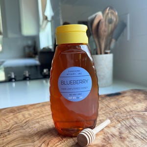 May include: A glass bottle of raw unfiltered blueberry honey with a yellow lid. The bottle has a white label with the text "Sunshine Apiary, Inc. Blueberry Raw Unfiltered Honey 561 602 1802 Net Wt 1/4 oz".