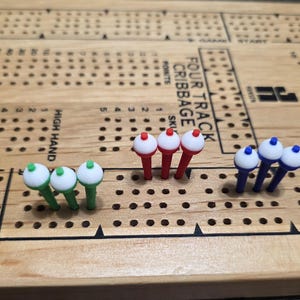 May include: A wooden cribbage board with four tracks, featuring multiple sets of colored pegs. The pegs are white-topped with green, red, and blue stems. The board has text that reads "FOUR TRACK CRIBBAGE" and "HIGH HAND".