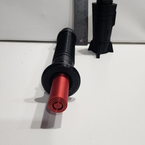 May include: A black and red 3D printed lightsaber prop with a red blade. The lightsaber is in two pieces, the blade and the hilt. The hilt has a textured surface and a black ring around the top.