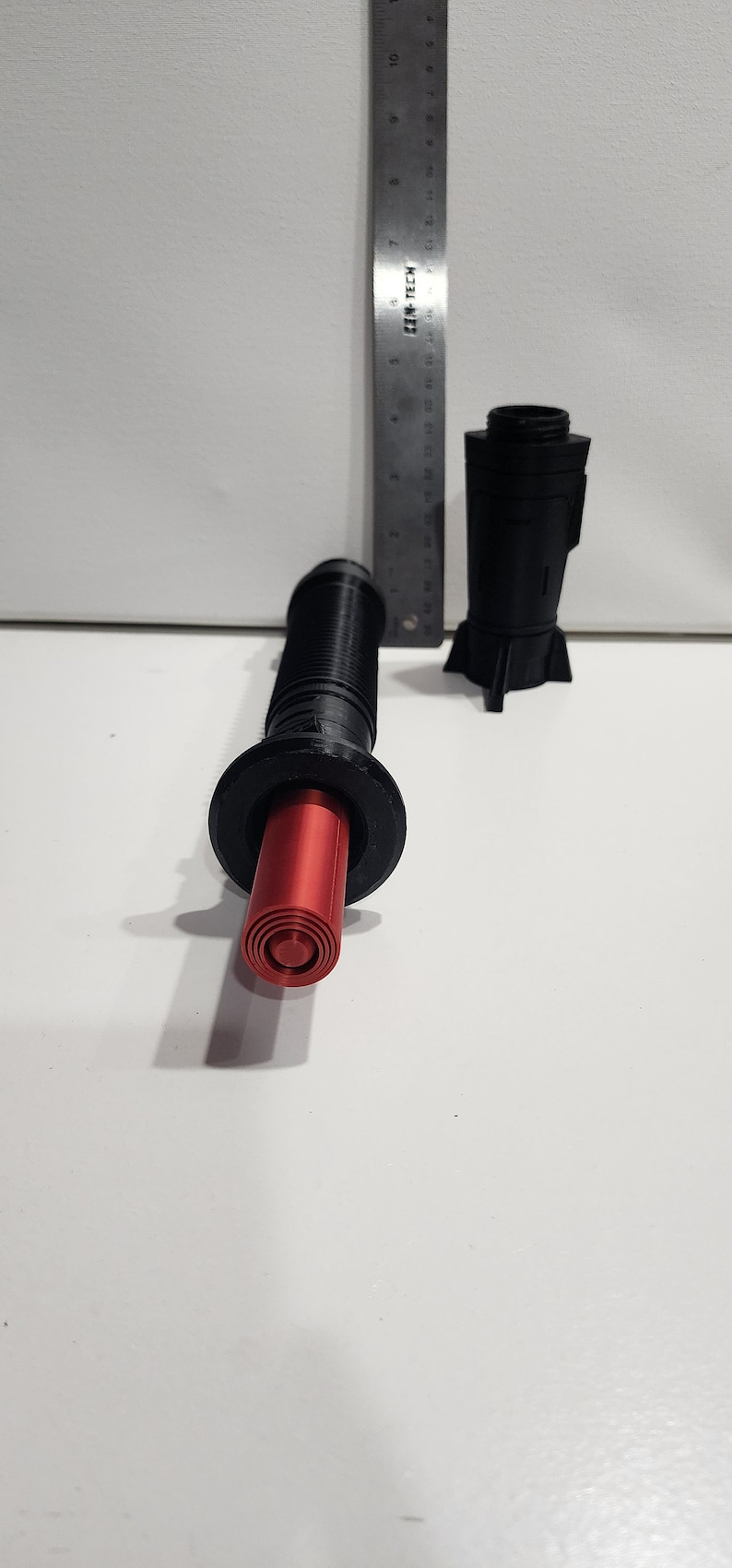May include: A black and red 3D printed lightsaber prop with a red blade. The lightsaber is in two pieces, the blade and the hilt. The hilt has a textured surface and a black ring around the top.