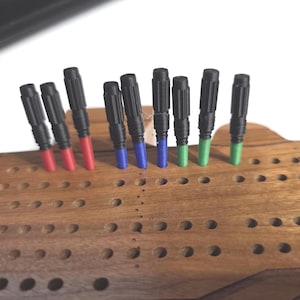 Pu&ograve; includere: Un tabellone di cribbage in legno con pioli neri e colorati. I pioli hanno la parte superiore nera e la parte inferiore colorata in rosso, blu e verde. Il tabellone &egrave; in legno e presenta diversi fori per i pioli.