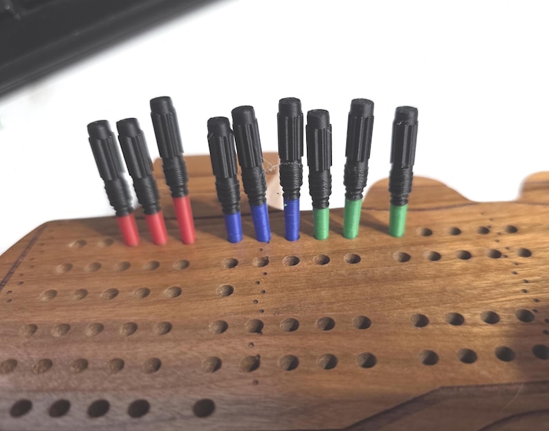 Pu&ograve; includere: Un tabellone di cribbage in legno con pioli neri e colorati. I pioli hanno la parte superiore nera e la parte inferiore colorata in rosso, blu e verde. Il tabellone &egrave; in legno e presenta diversi fori per i pioli.