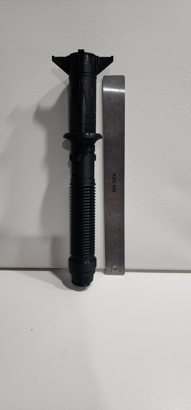 May include: A black 3D printed lightsaber prop, approximately 11 inches long, with a ribbed handle and a detailed hilt. The prop is next to a ruler for scale.