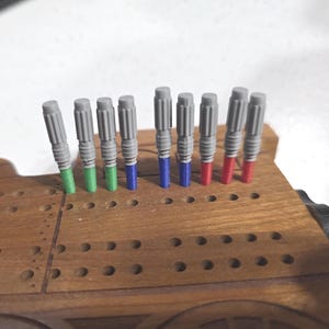 May include: A wooden cribbage board with a train design, featuring a series of holes for peg placement. The board holds gray pegs with green, blue, and red tips. The pegs are inserted into the board's holes.