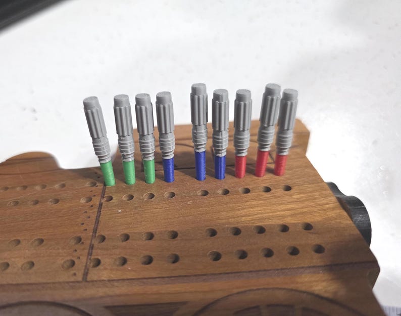 Pu&ograve; includere: Un gioco da tavolo di cribbage in legno a forma di treno, con fori per i pioli. Il gioco contiene pioli grigi con punte verdi, blu e rosse. I pioli sono inseriti nei fori del tabellone.