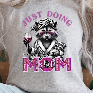 May include: Gray sweatshirt with a cartoon raccoon wearing sunglasses and a robe, holding a glass of wine. The text "Just Doing Mom Shit" is written around the raccoon in pink.