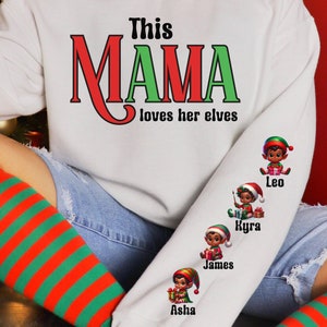 May include: White sweatshirt with the text "This MAMA loves her elves" in red and green. The sleeve features elf illustrations with names: Leo, Kyra, James, and Asha. The person is wearing red and green striped socks.