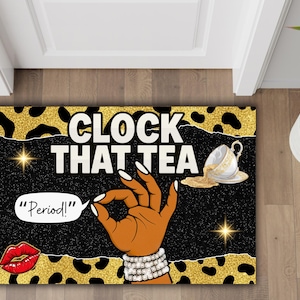 May include: Door mat with a black background and gold leopard print border. The mat features the text "CLOCK THAT TEA" and a hand gesture. A spilled teacup and red lips are also shown.