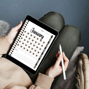 May include: A person is holding a tablet with a digital calendar open to January. The calendar has brown squares for each day. The person is holding a white stylus in their other hand.