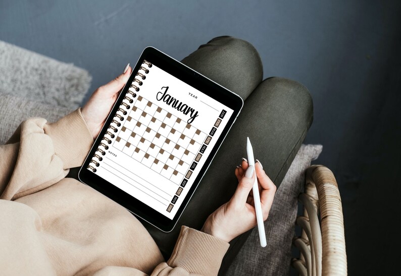 May include: A person is holding a tablet with a digital calendar open to January. The calendar has brown squares for each day. The person is holding a white stylus in their other hand.