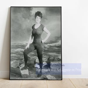 May include: Black and white framed print of a woman in a vintage swimsuit posing on a rock. The one-piece swimsuit has short sleeves and is paired with leggings. The background shows a cloudy sky and ocean waves.