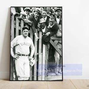 May include: Black and white photo of a baseball player wearing a "YANKEE" jersey, standing in front of a group of men. The player is holding a baseball bat and a baseball glove.