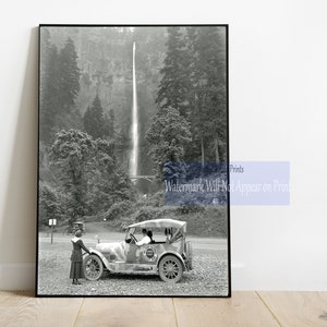 May include: A black and white photograph of a vintage car parked in front of a waterfall. The car is a touring car with a canvas top and a spare tire mounted on the back. There are two people in the car, and a woman standing next to the car.