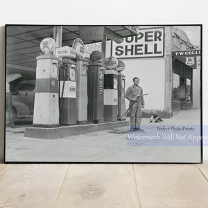 May include: A black and white photo of a gas station with four pumps. The pumps are labeled "PLUME" and "SUPER SHELL". A man in a light-colored shirt and pants stands next to the pumps, holding a hose. A dog sits on the ground in the foreground.