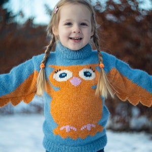 May include: A young girl wearing a blue and orange knitted jumper with an owl design. The jumper has a high neck and long sleeves. The girl has blonde hair in plaits and is smiling.