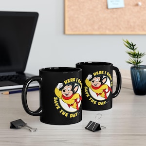 May include: Two black ceramic mugs with a cartoon mouse superhero design. The mouse is wearing a yellow outfit and red cape, with the text "HERE I COME TO SAVE THE DAY" in a circular design.