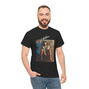 Puede incluir: Una camiseta negra de manga corta con un estampado gráfico de una persona con un top gris de hombros descubiertos y tacones rojos sentada en una silla. La palabra Flashdance está impresa en blanco sobre la imagen con líneas azules.