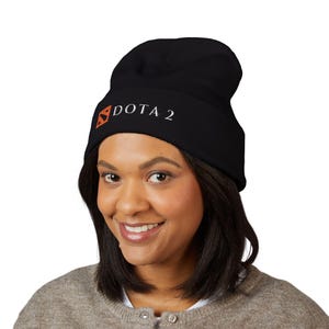 Puede incluir: Gorro negro con el texto "DOTA 2" en blanco y un logotipo naranja. El gorro está hecho de un material suave y está diseñado para mantener la cabeza caliente. Un accesorio popular para los jugadores.
