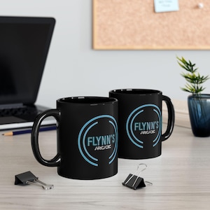 Puede incluir: Dos tazas de café negras con el texto "FLYNN'S ARCADE" en un diseño circular turquesa y blanco. Las tazas están sobre una superficie de color claro, con un portátil y material de oficina en el fondo.