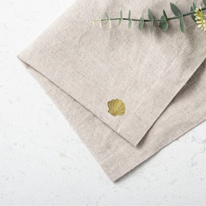 Puede incluir: Una servilleta de lino beige con una concha dorada bordada en el centro.