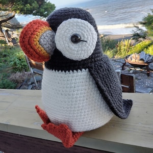 May include: A handmade crochet puffin plush toy featuring a dark gray back and wings, a white belly, and a white face patch. It has a prominent, multi-colored beak in shades of orange, yellow, and gray, orange feet, and a single black button eye.