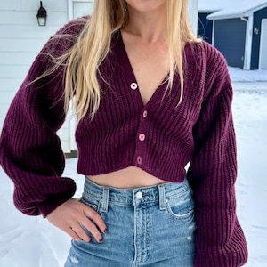 May include: A plum-coloured, cropped cardigan with a V-neck and bubble sleeves. The sweater has a ribbed knit texture and is fastened with pink buttons. It is paired with light-wash blue denim jeans.